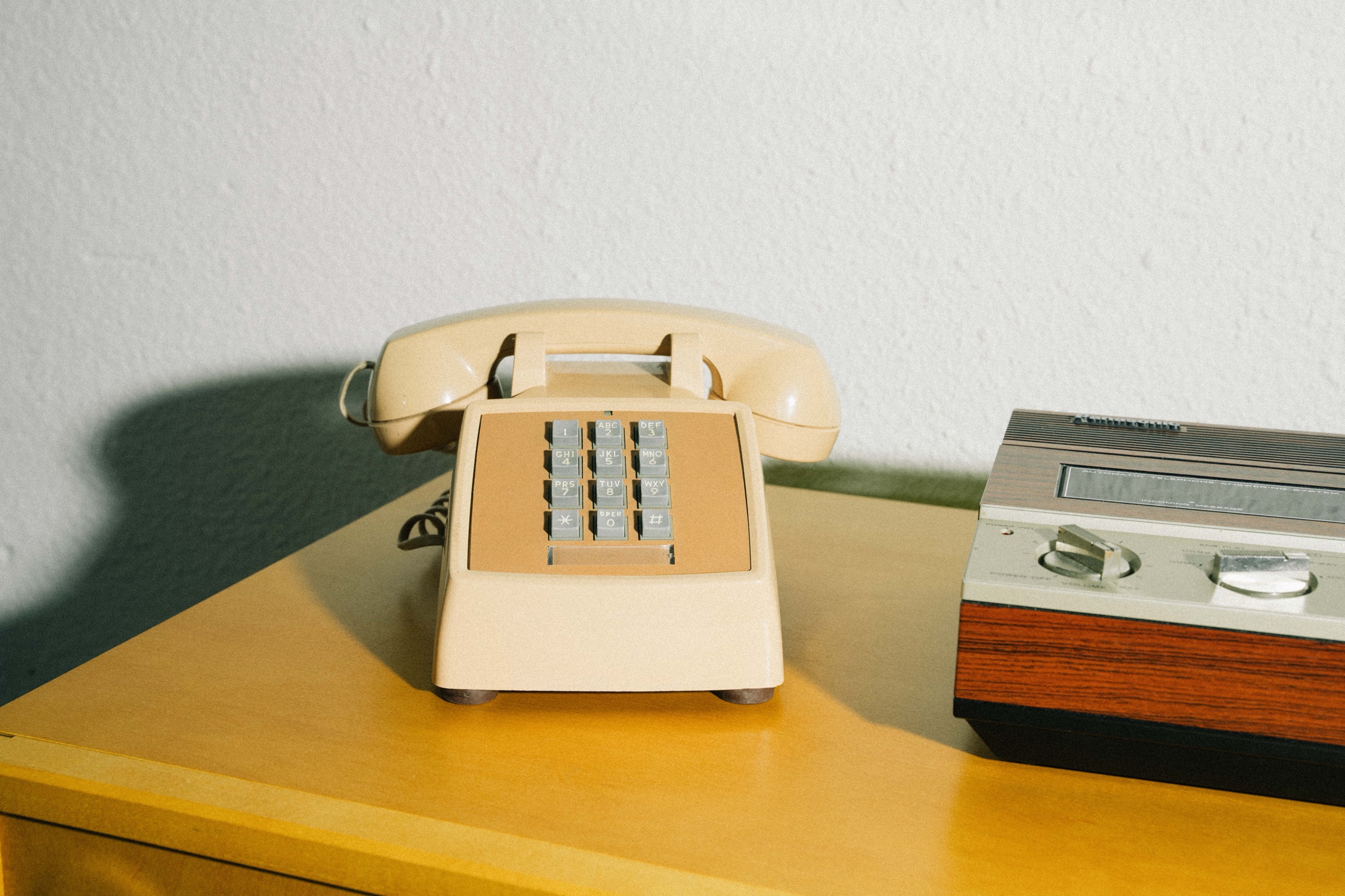 A cream push-button desk phone and a wood-grain answering machine on a yellow cabinet in warm sunlight.