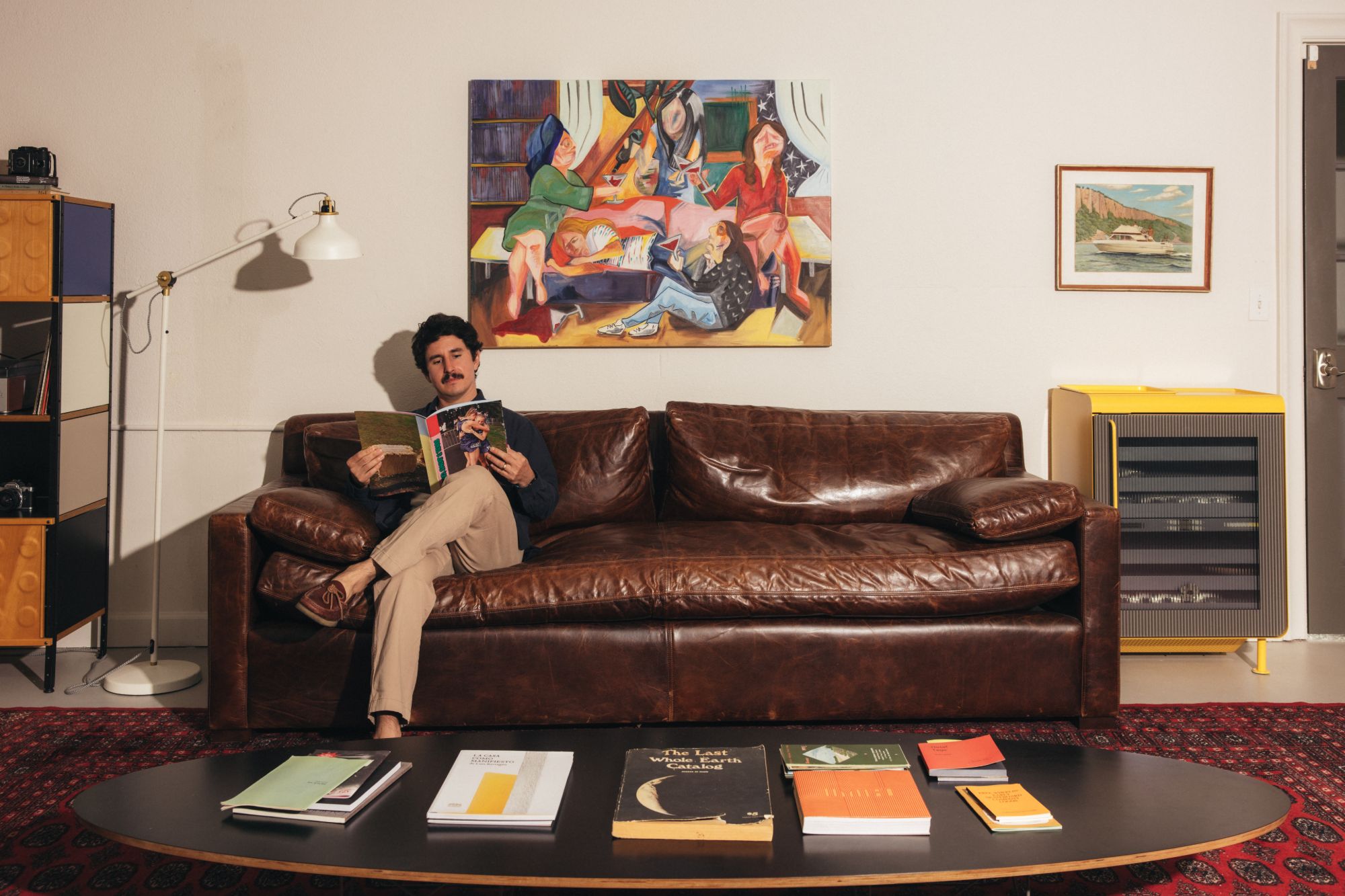 A person reading on the leather sofa beneath the painting, with a stack of books laid out on the coffee table in the foreground.
