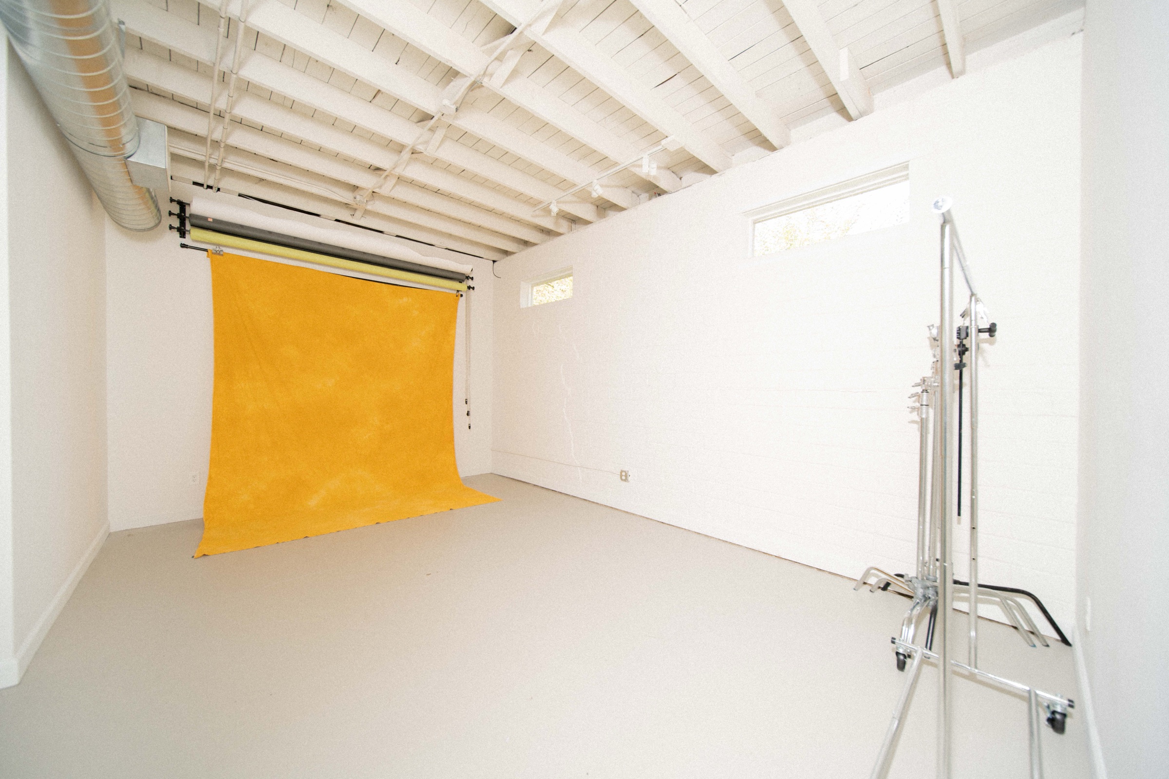 The Ace Studio shooting room: white painted brick walls and exposed ceiling beams, seamless paper rolls with a yellow muslin backdrop hung, and a C-stand at right.